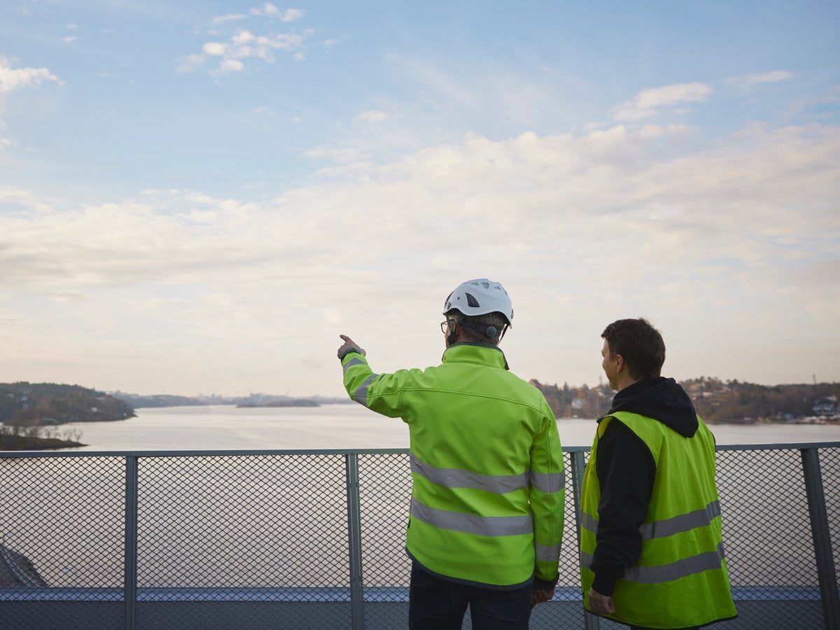 Två personer i reflexkläder, den ena med bygghjälm, tittar ut över vattnet. En person pekar bortåt.