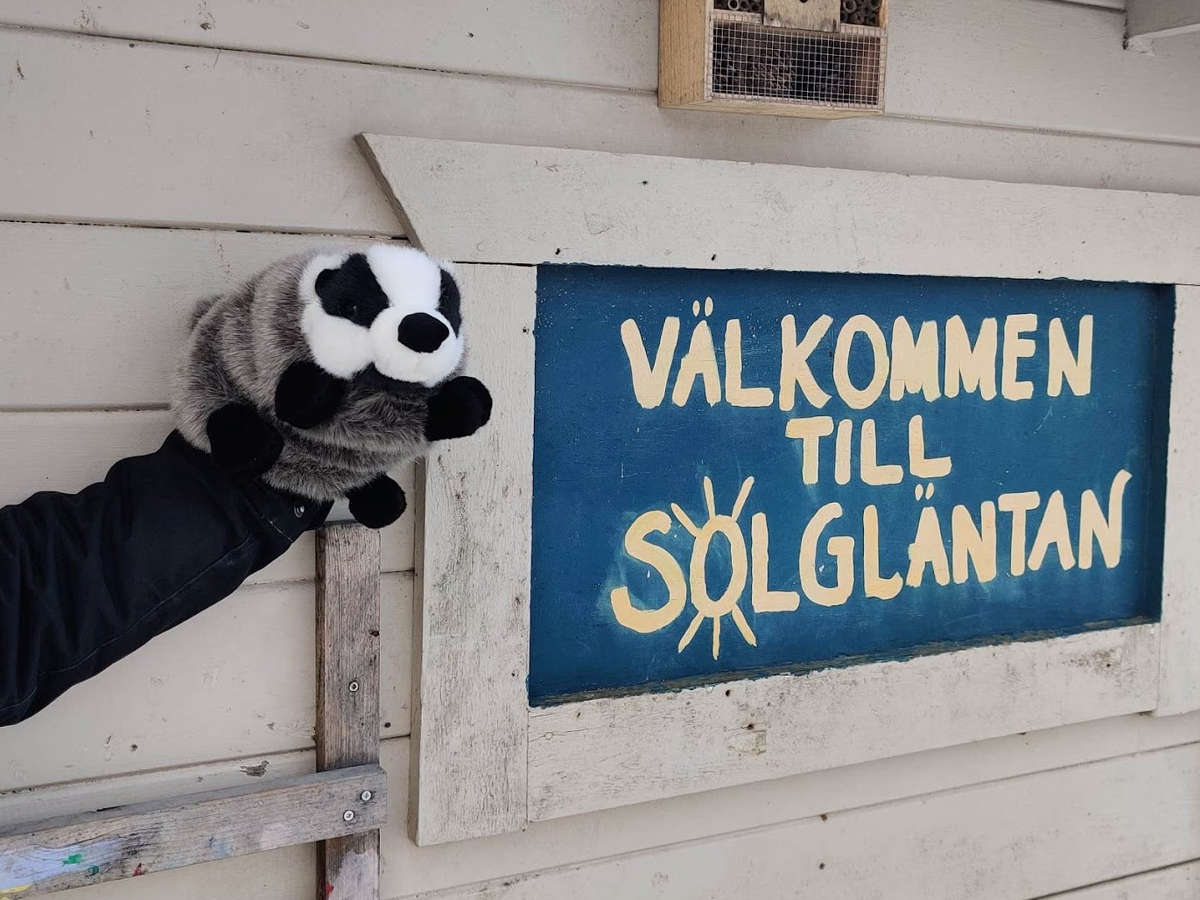 En person håller en handdocka med en grävling bredvid en skylt där det står. Välkommen till Solgläntan.