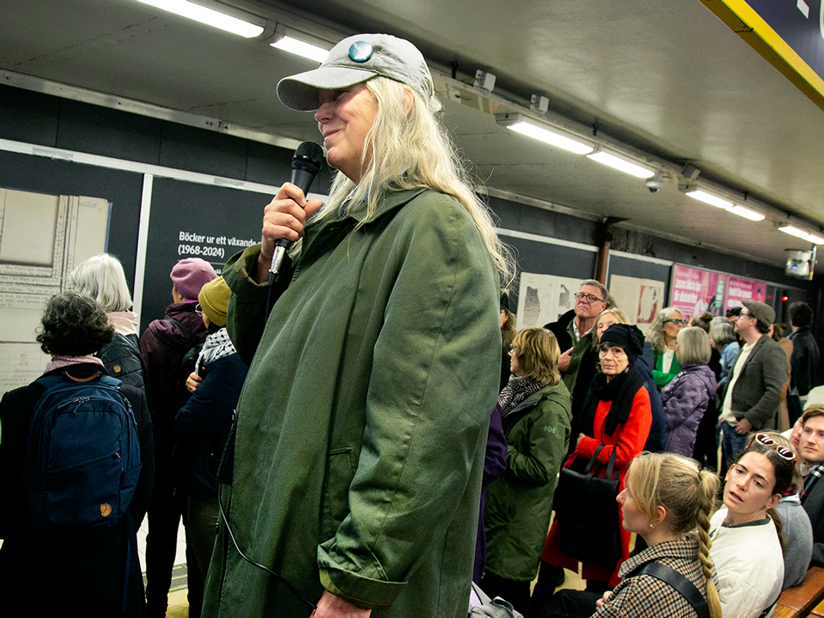 Marja-leena i keps står på bänk på Slussens tunnelbanestation. Många människor runt om kring som tittar.