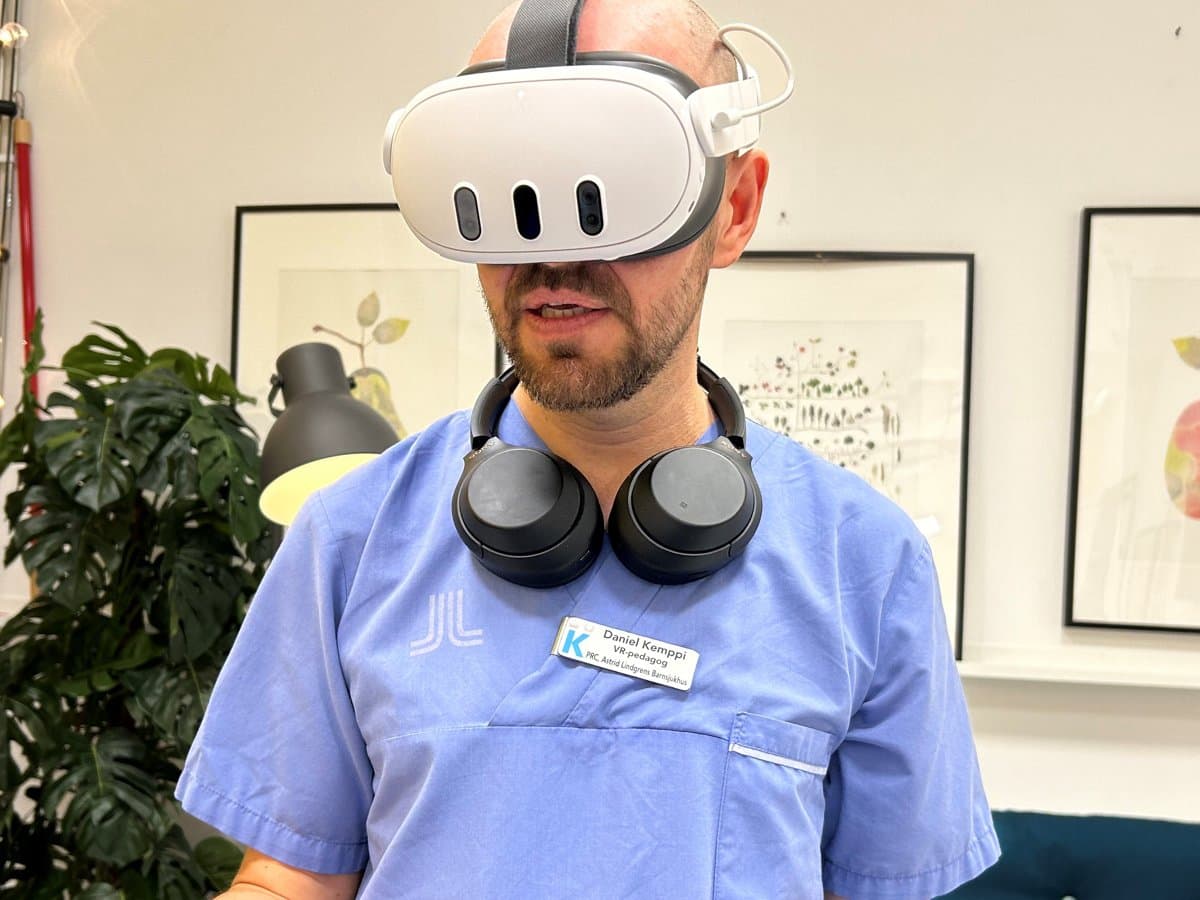 En man med blå sjukhusskjorta står med vr-headset på huvudet och handkontroller i handen
