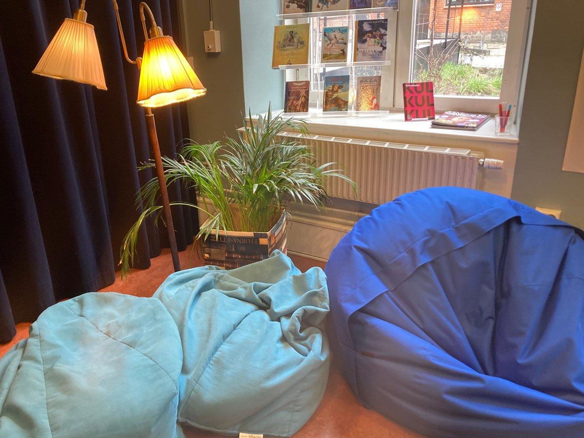 Bild på läshörna på Hornstulls bibliotek. Blåa sittpuffar, en krukväxt och en golvlampa framför ett fönster med böcker. 