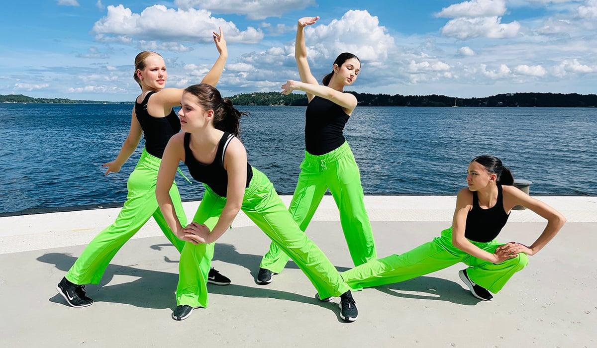 Fyra flickor i gröna byxor och svarta linnen dansar å en brygga vid havet.