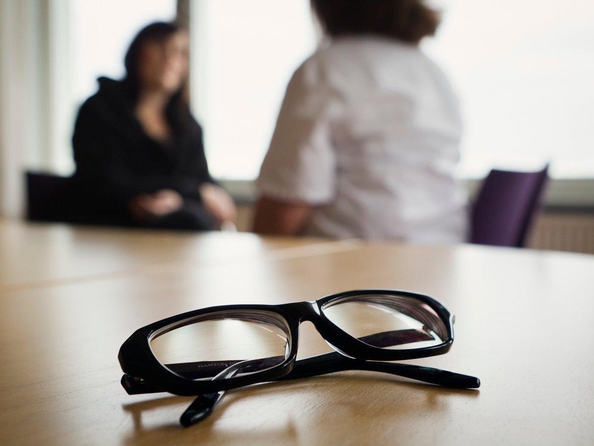 Närbild på glasögon som ligger på ett bord. I bakgrunden syns två personer sitta och prata med varandra. 