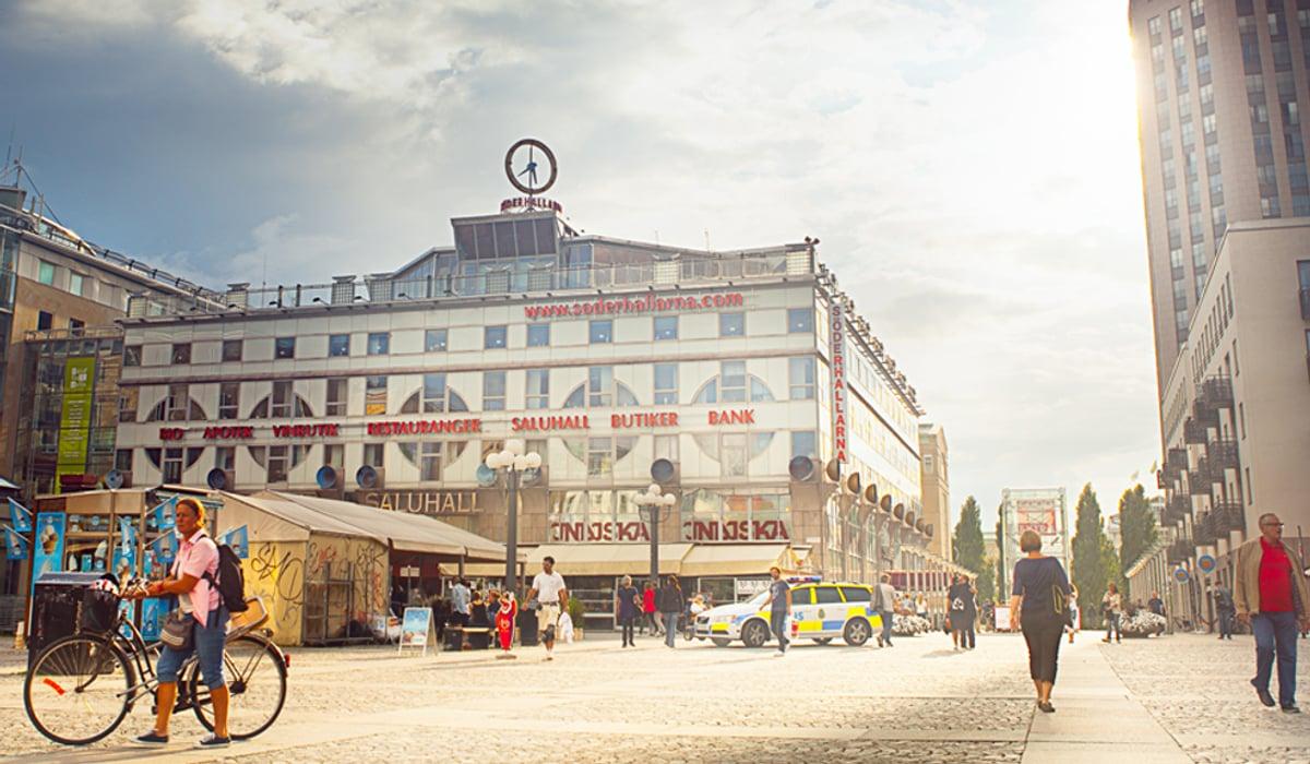 Foto över Medborgarplatsen i Stockholm. I bilden är det soligt sommarväder och personer som går över torget.