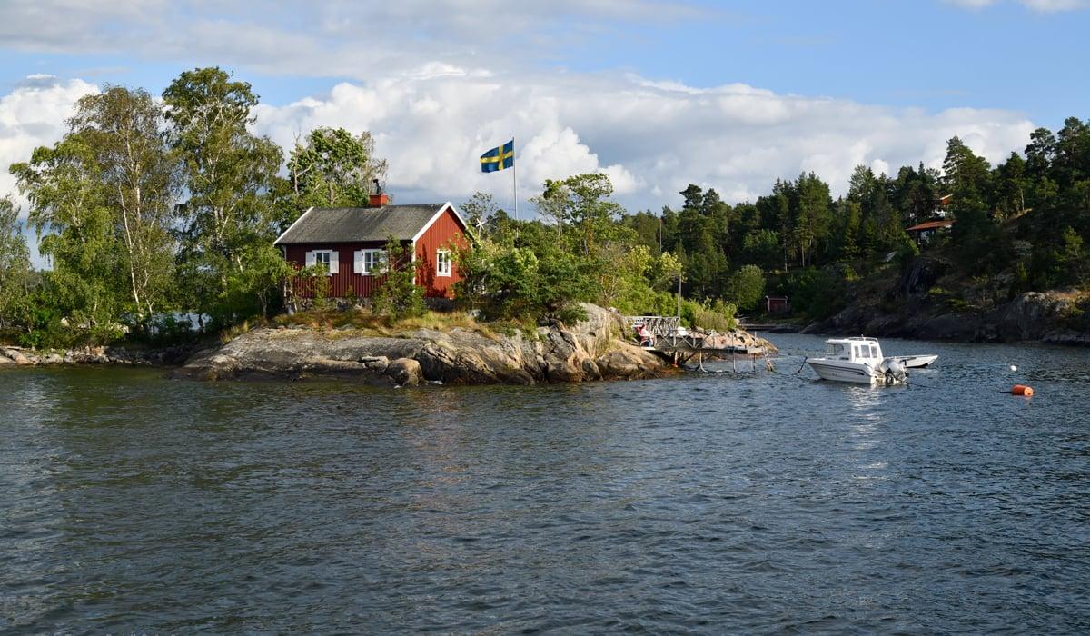Bild på liten skärgårdsö med rött hus med vita knutar och svensk flagga som svajar i flaggstång. 