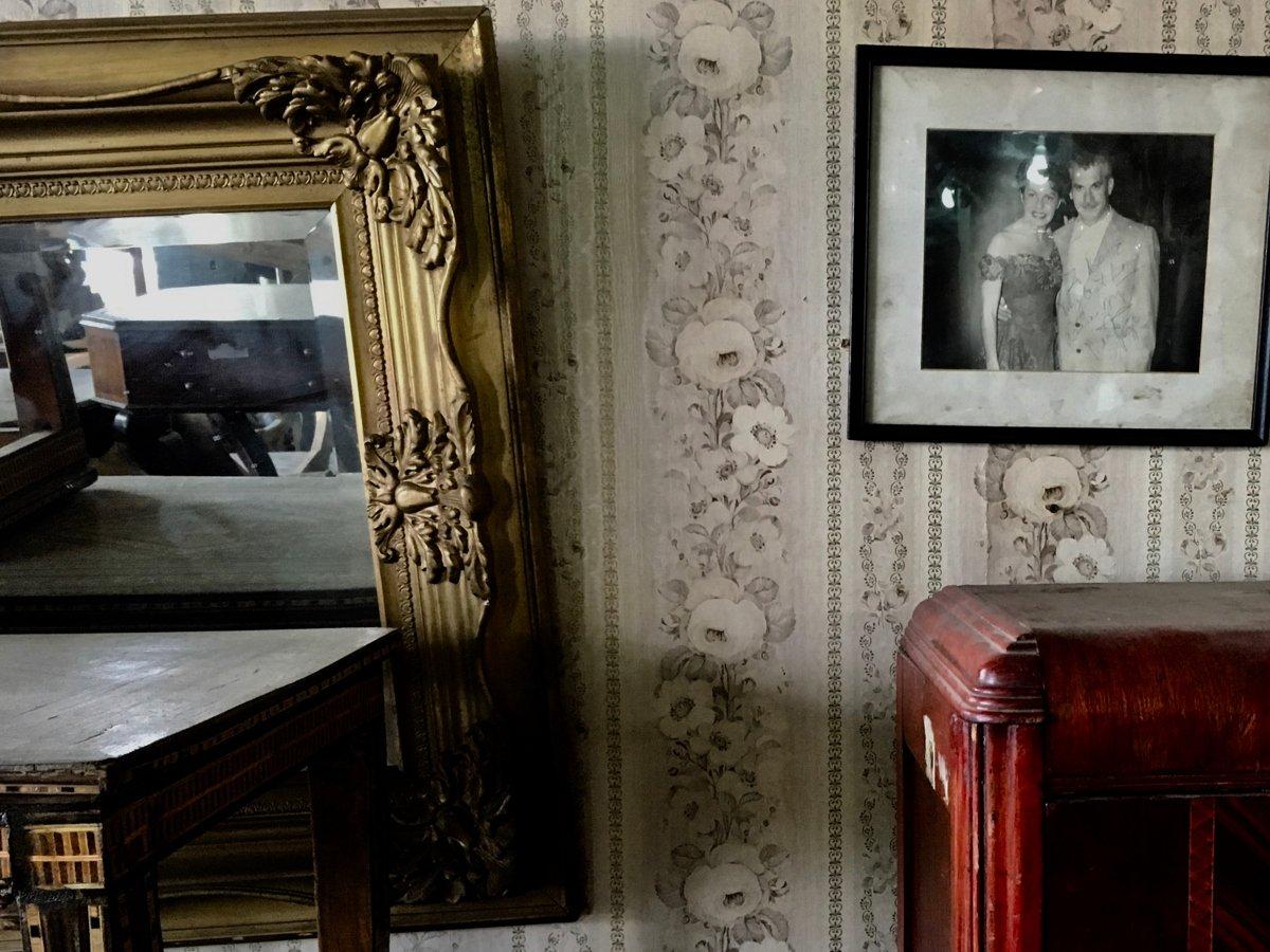 Stillbild från film. Dammigt bord och skåp under en spegel med guldram och ett fotografi som hänger snett på väggen.