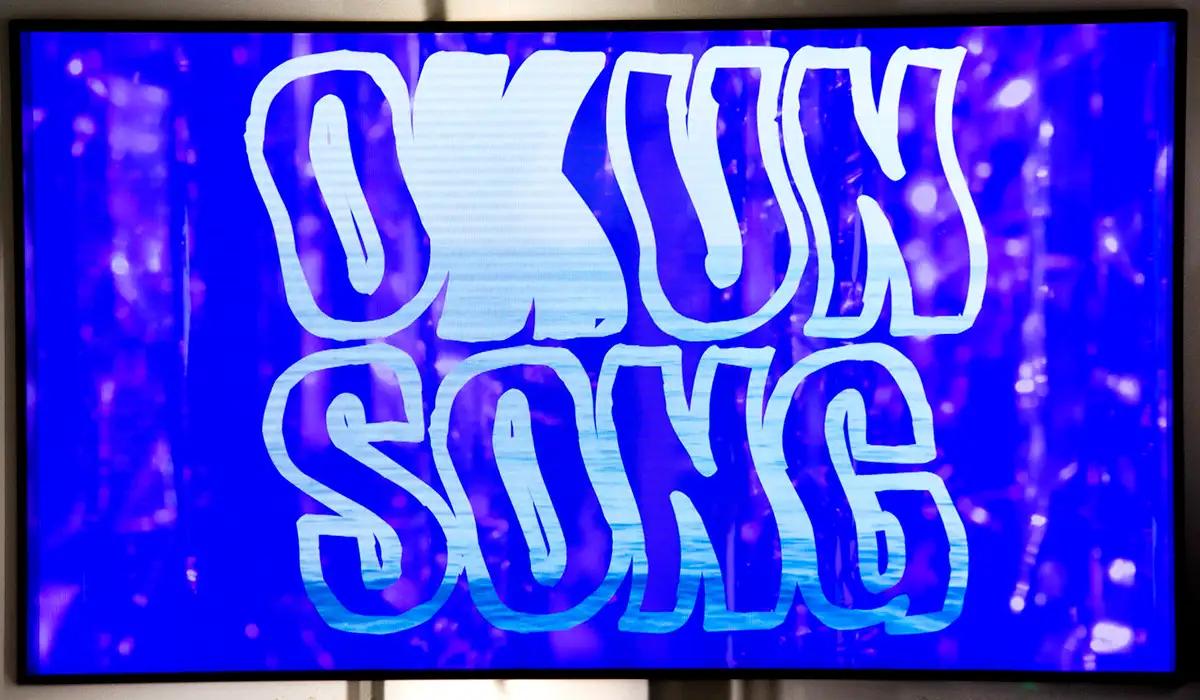 Texten Okun Song på blå bakgrund på en videoskärm.