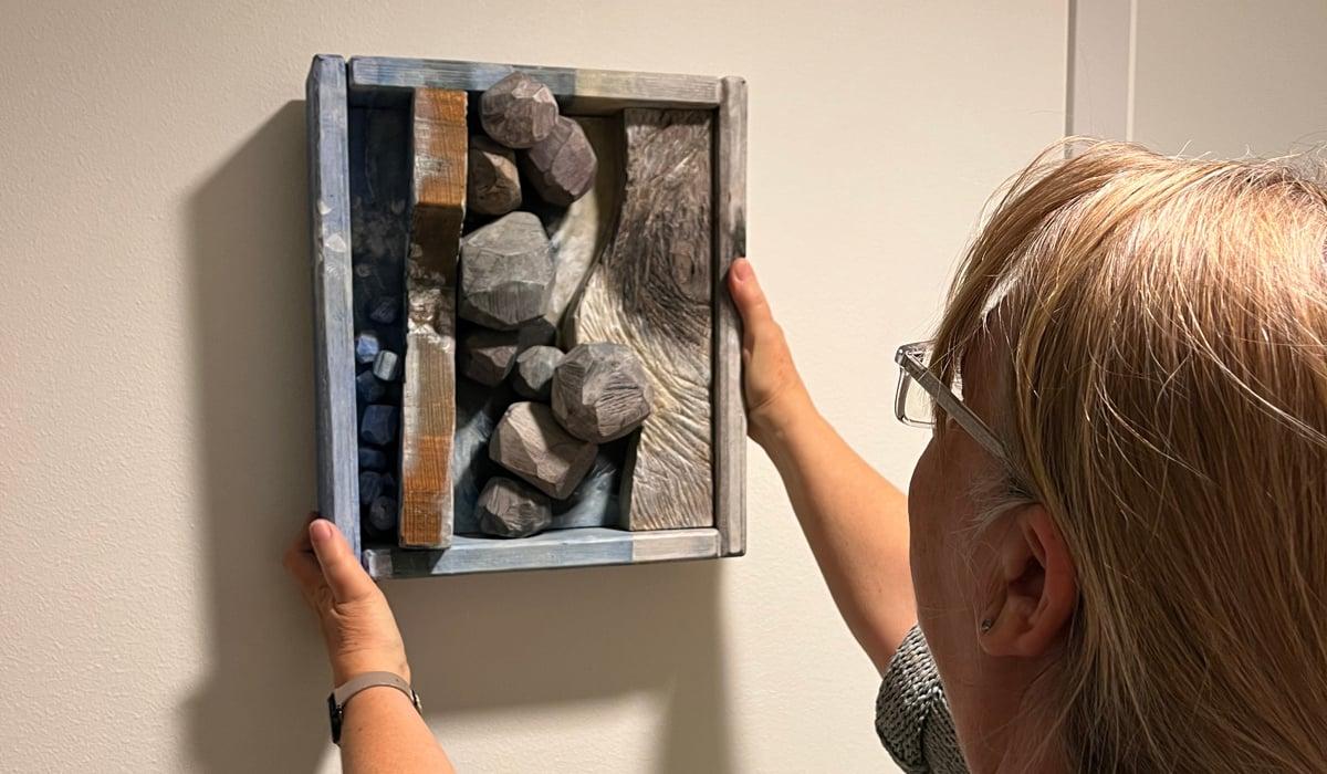 Kvinna hänger upp en tavla på väggen. Det är en låda med stenar och trä monterade sida vid sida.