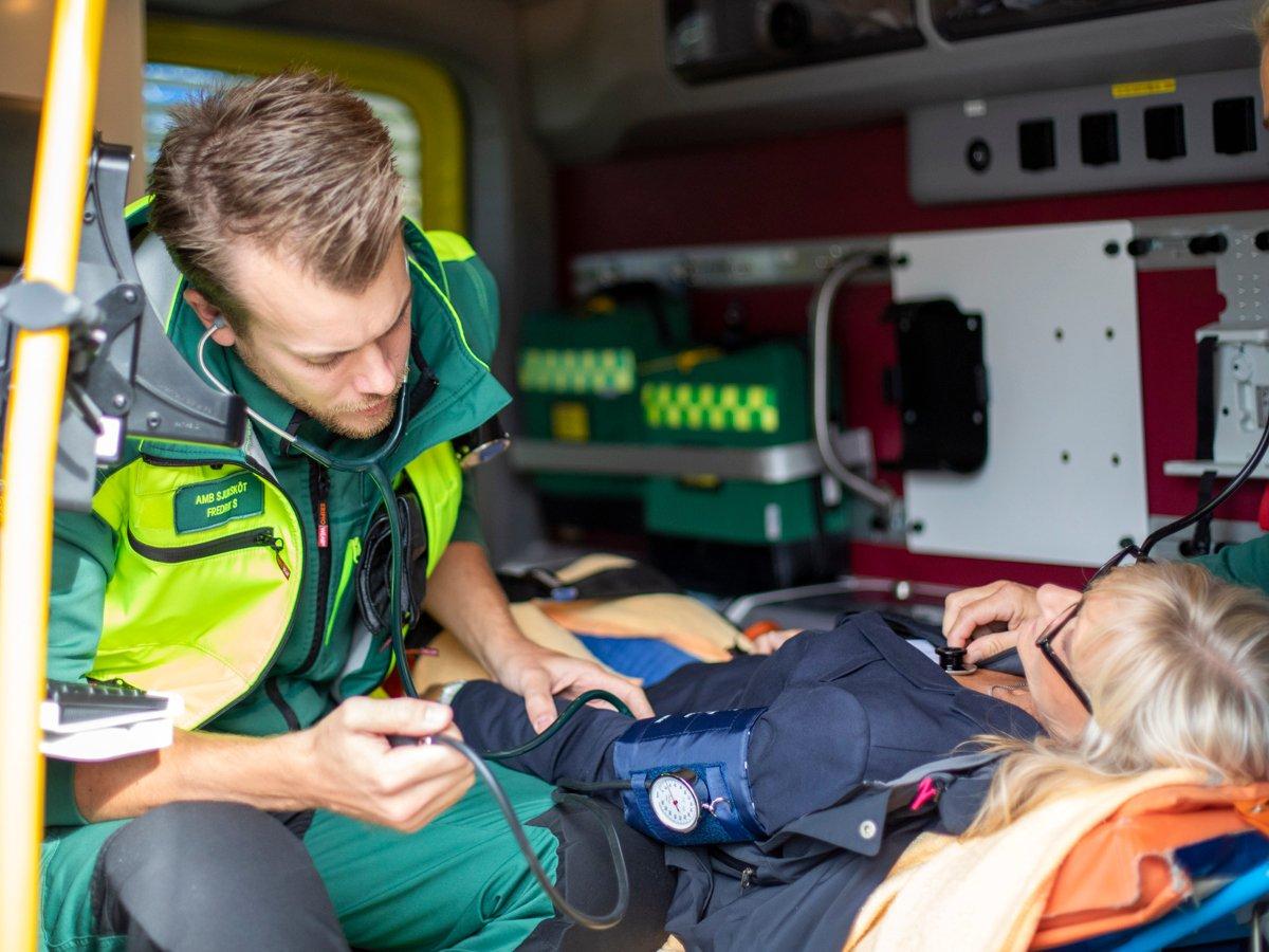 Två medarbetare inom ambulanssjukvården i neongröna arbetskläder tar hand om en kvinnlig patient som sitter på en bår.