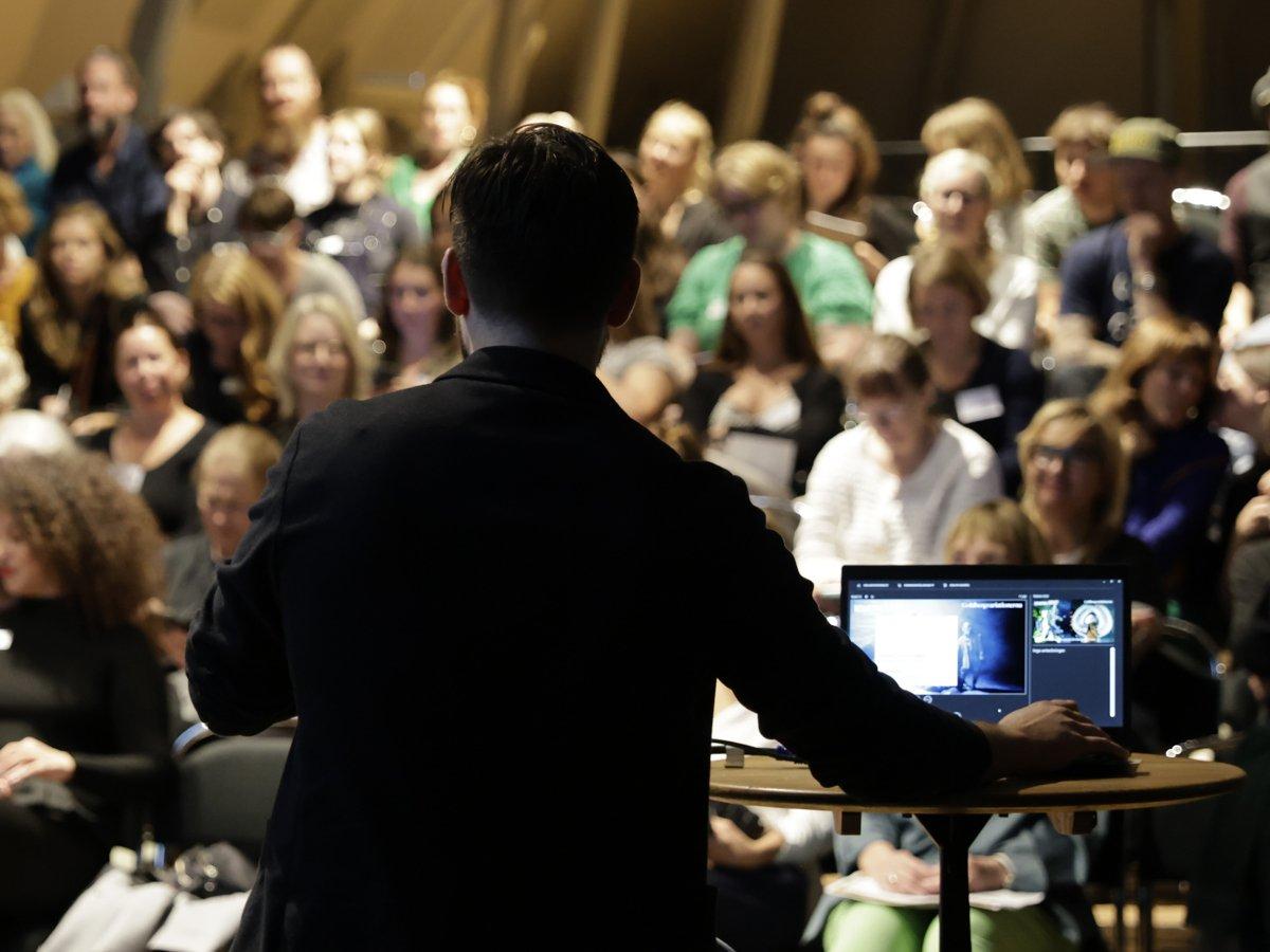 En lokal fullsatt med publik, med ryggen på en föreläsare i förgrunden