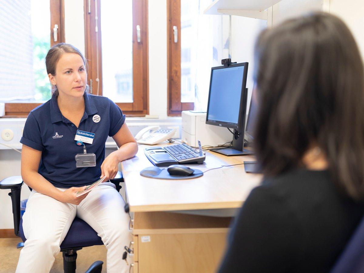 Sjuksköterska på vårdcentral sitter vid ett skrivbord och pratar med en patient som sitter mittemot.