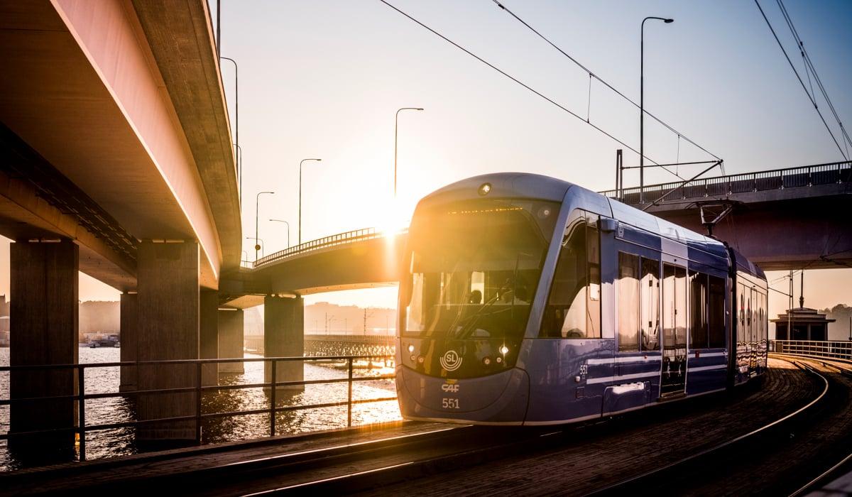 Lidingöbanans tåg rullar mellan lidingöbroarna. Det är en stark solnedgång som skiner över området.