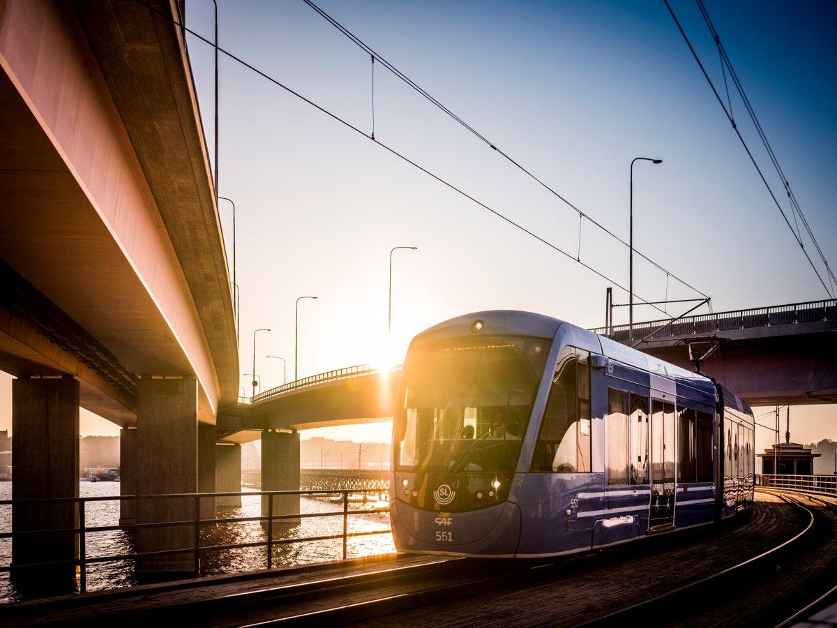 Lidingöbanans tåg rullar mellan lidingöbroarna. Det är en stark solnedgång som skiner över området.