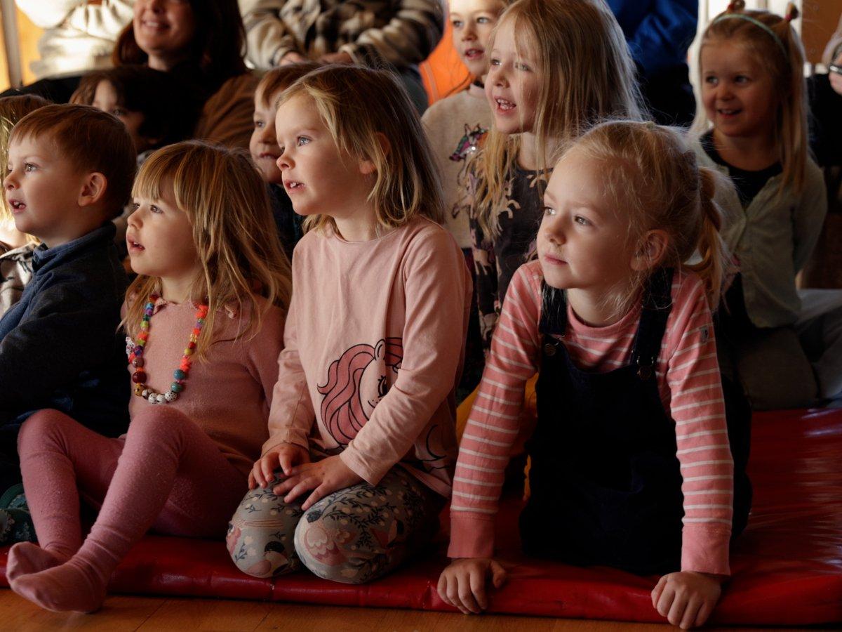 Barn som sitter på dynor på ett golv och tittar koncentrerat åt samma håll (föreställningen de tittar på är utanför bilden)