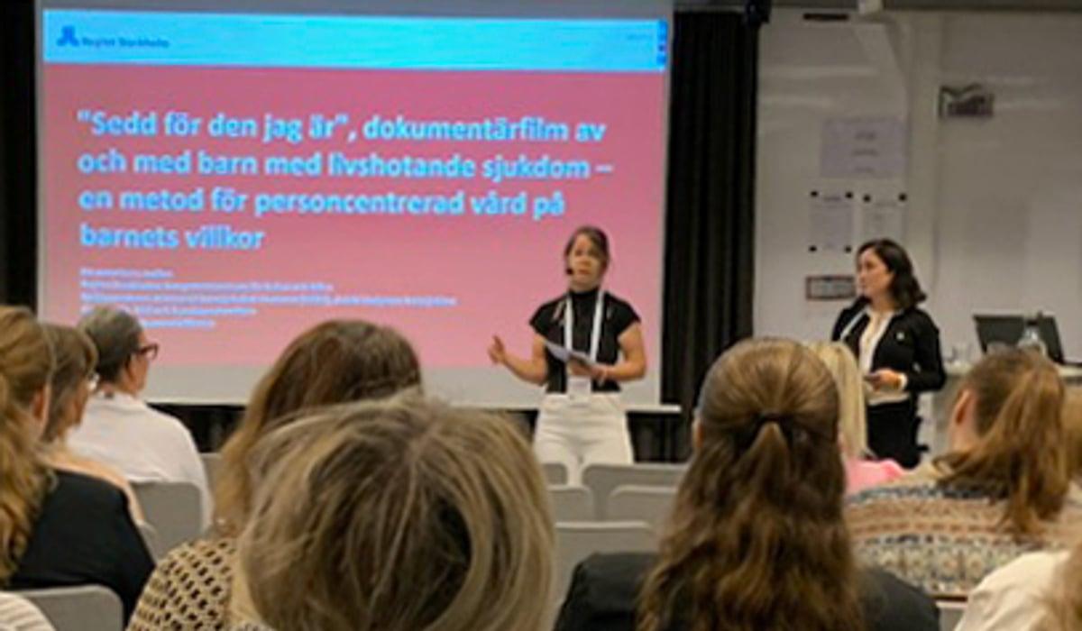 Konferensdeltagare tittar på en skärm och två talare