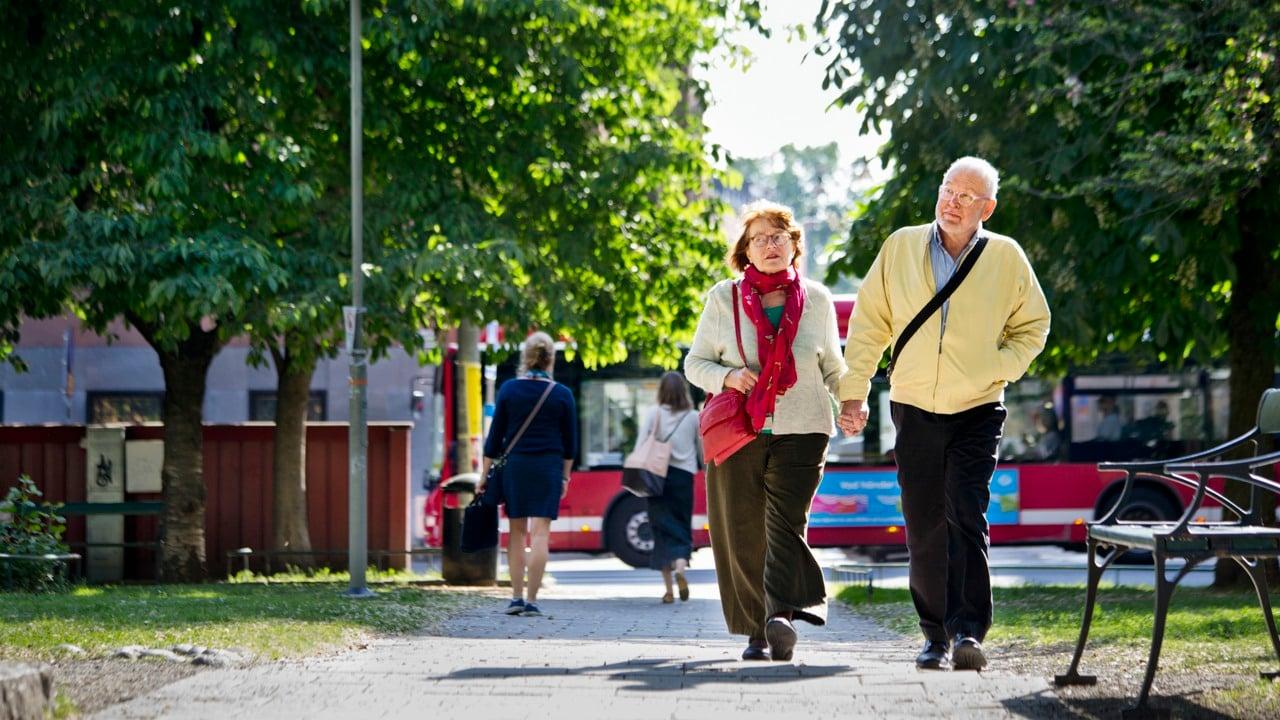 Två äldre som går på promenad i en lummig park. De håller hand. 