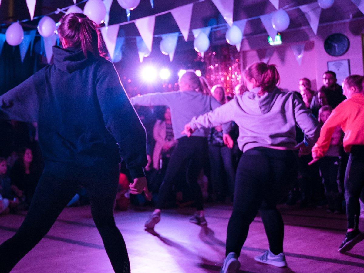 Fem dansare dansar en synkroniserad koreografi på ett dansgolv i en mörk lokal med flaggor, ballonger och festlig belysning i taket.