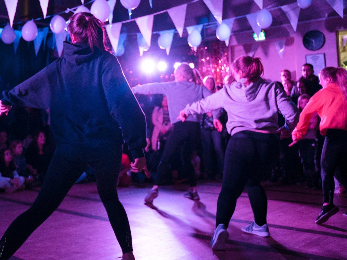 Fem dansare dansar en synkroniserad koreografi på ett dansgolv i en mörk lokal med flaggor, ballonger och festlig belysning i taket.