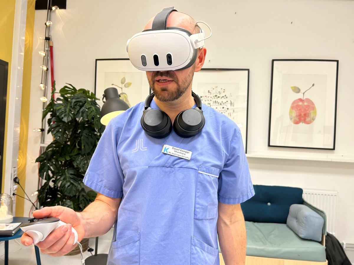 En man med blå sjukhusskjorta står med vr-headset på huvudet och handkontroller i handen
