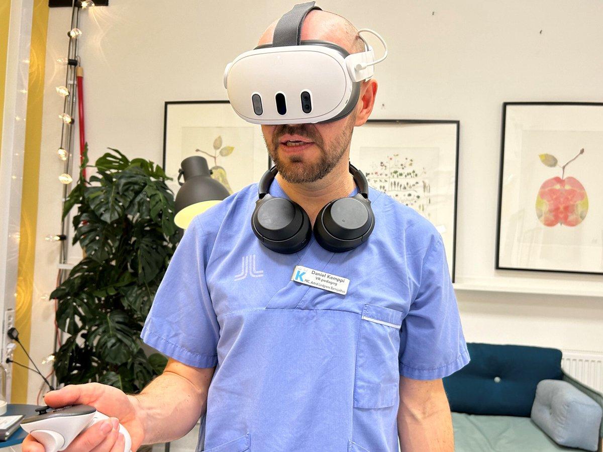 En man med blå sjukhusskjorta står med vr-headset på huvudet och handkontroller i handen
