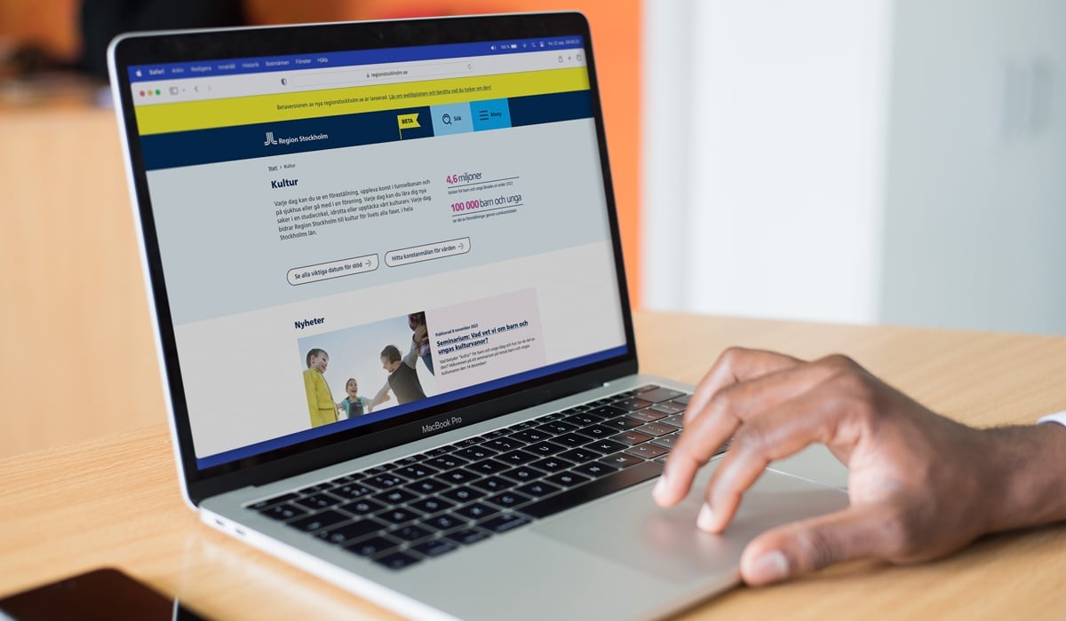 Foto på en laptop som står på ett bord. En person som inte syns i bild har sin hand på musplattan. På skärmen är Region Stockholms testversion av sin nya webbsida öppen. Till vänster om datorn ligger en mobiltelefon på bordet.