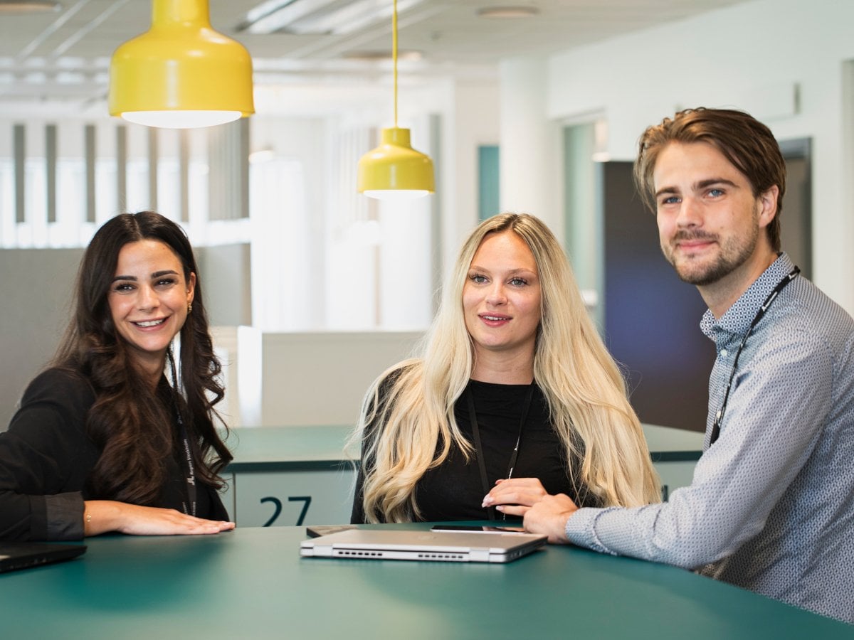 Tre unga kollegor står i ett kontorslandskap vid ett grönt bord och samtalar.