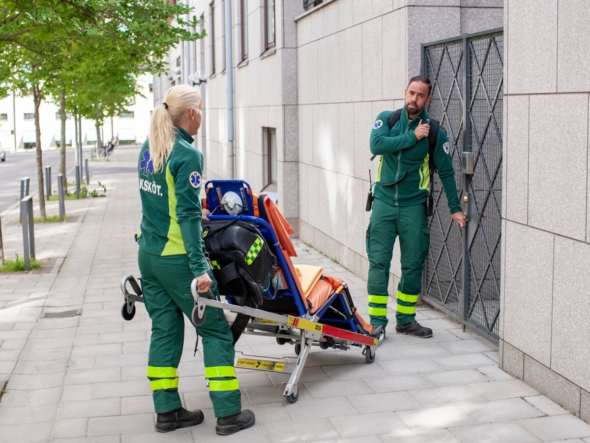 Två medarbetare i gröna arbetskläder väntar på att bli insläppa genom en grind. Den kvinnliga medarbetaren håller i en bår